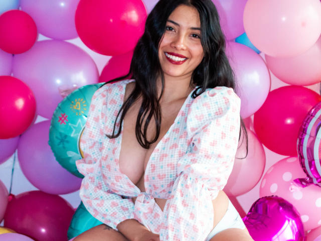 IsabellaPrincess with long black hair, smiling, wearing a tied-up, checkered shirt exposing her big tits, and white panties, surrounded by pink and purple balloons.