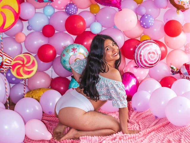 IsabellaPrincess, con lunghi capelli neri, sorridente, inginocchiata, indossa una maglietta a spalle scoperte con motivi floreali e biancheria intima bianca, ed è circondata da colorati palloncini e decorazioni a tema dolci. Il suo sedere è ben visibile..