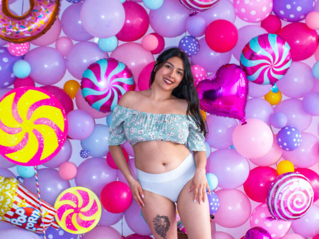 Isabella: Principessa con lunghi capelli neri, top floreale con spalline scoperte, biancheria intima bianca, che sorride, in piedi in mezzo a palloncini colorati, decorazioni a tema cuore e caramelle, tatuaggio visibile sulla coscia destra..