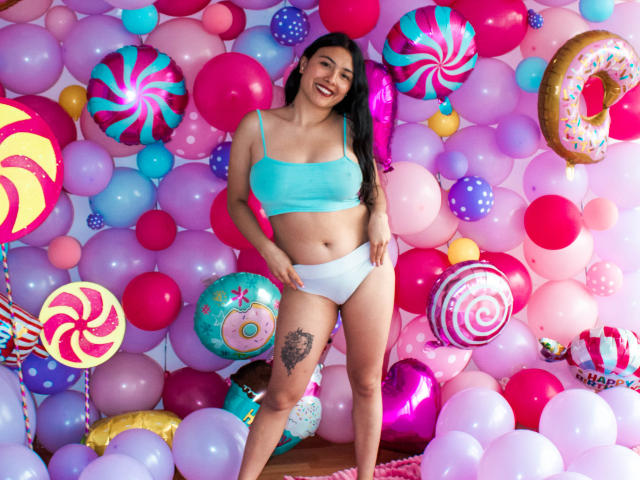 Isabella, una principessa con lunghi capelli neri, sorridente, che indossa un reggiseno turchese e biancheria intima bianca, in piedi di fronte a palloncini colorati, che mostra un tatuaggio sulla coscia e un corpo carino e formoso..