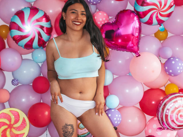 IsabellaPrincess with long black hair, smiling, wearing a light blue crop top and white panties, standing among colorful balloons, showing a tattoo on her right thigh.