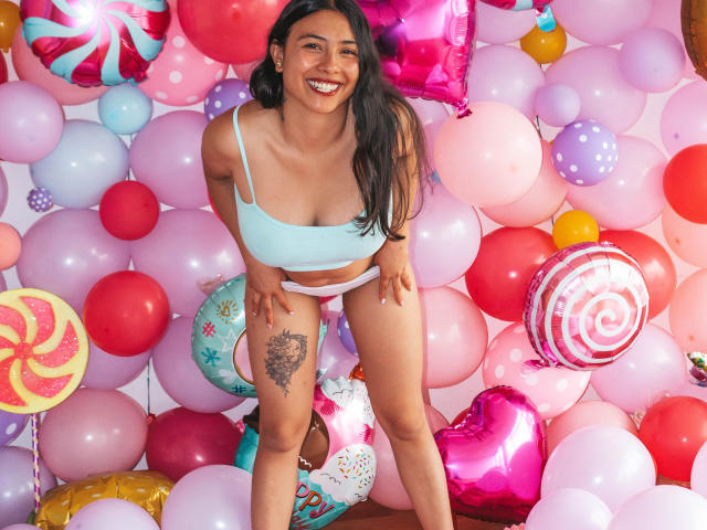 Isabella, una principessa con lunghi capelli neri, sorridente, che indossa un top corto azzurro chiaro, slip bianchi e rossi a righe, piegata in avanti, mostrando un tatuaggio sulla coscia destra, circondata da palloncini..