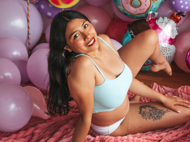 IsabellaPrincess, Latina with long black hair, smiling, wearing a light blue sports bra and white panties, sitting on pink blanket, showing a tattoo on her right thigh.