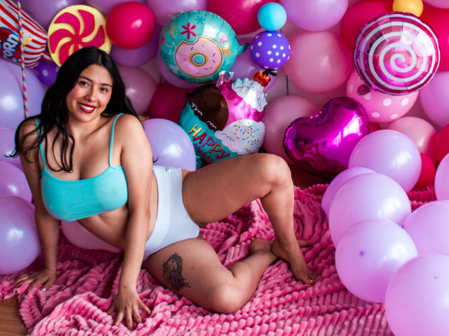 Isabella, una principessa formosa, con un reggiseno blu e biancheria intima bianca, seduta su una coperta rosa, circondata da palloncini colorati e decorazioni a tema dolci, sorride alla fotocamera..