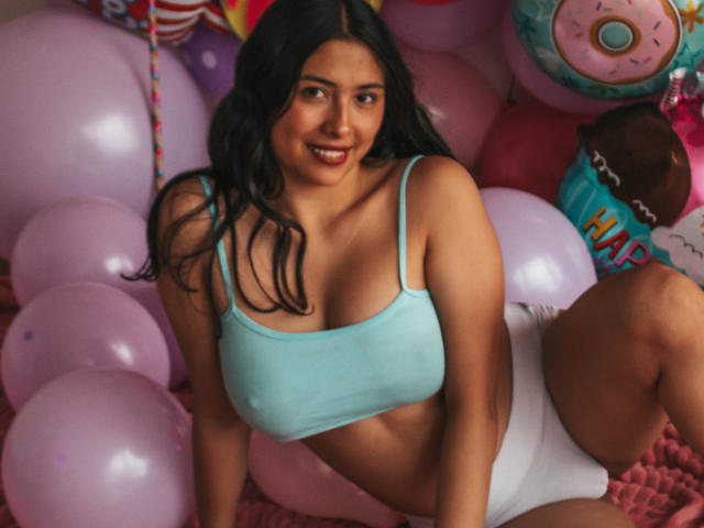 IsabellaPrincess with dark wavy hair, smiling, wearing a light blue crop top showing her tits and white shorts, surrounded by pink balloons, sitting on a pink rug.