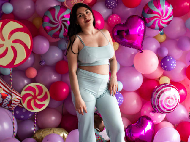 IsabellaPrincess with medium tits, wearing a light blue crop top and matching pants, standing in a colorful balloon wall with candy-themed shapes. She has long dark hair and red lipstick.