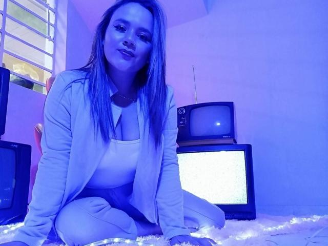 NatalieRey, in blauwige tinten, zittend op een pluizig tapijt, gekleed in een witte top en een vest, met lang, steil haar, kijkend naar de camera, omringd door oude televisies..