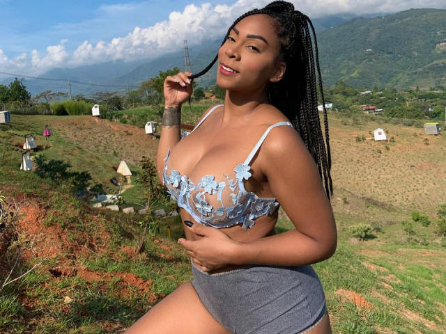 EbonyEnith with braided hair, blue lace bra, showing her tits, gray shorts, smiling, holding a braid, outdoors, mountains, clear sky, tattoo on right forearm.