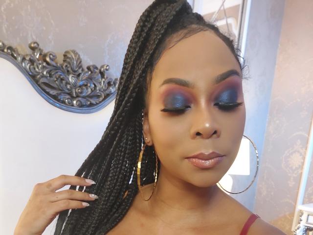 EbonyEnith with dark braids, blue eyeshadow, and pink lipstick, wearing gold hoop earrings, has glossy lips and a calm expression, touching her braided hair with her left hand.