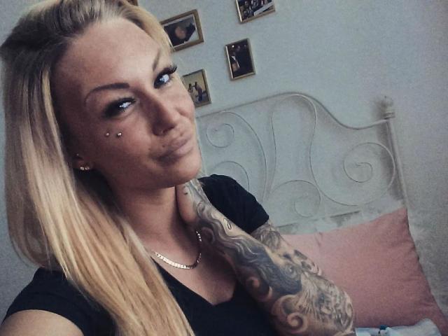 LadyXenia-hot with blonde hair, black top, tattoos on right arm, piercings on left cheek, smiling, holding neck, white metal bed, pink pillow, necklace.