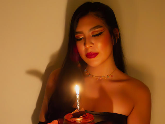 SamanthaEva69, com cabelo escuro, maquiagem vibrante, batom vermelho, segurando uma vela acesa, um pequeno colar de ouro, blusa preta sem alças, sombra na parede, olhando para baixo, luz de vela em sua pele..