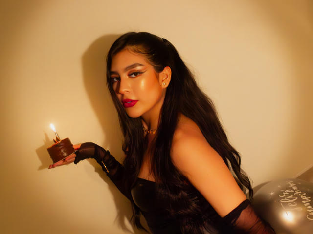 SamanthaEva69, con cabello negro largo, lápiz labial rojo y vestido negro, sosteniendo un pastel de chocolate con velas en su mano enguantada de negro, mirando seductoramente a la cámara..