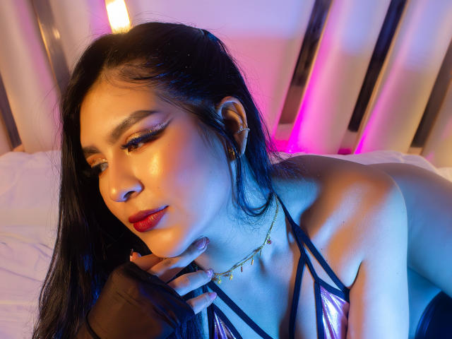 SamanthaEva69 with long black hair, red lipstick, shiny skin, gold necklace, and strappy metallic bra, lying on bed, looking down with hand on chin, blue and pink lights.