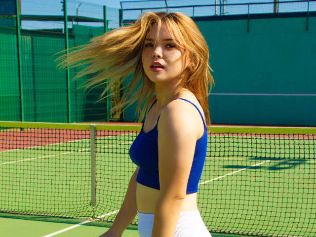 KiraAltman with blonde hair, blue sports bra, and white pants, standing on a tennis court, looking at the camera, her tits slightly visible, wind blowing through her hair.