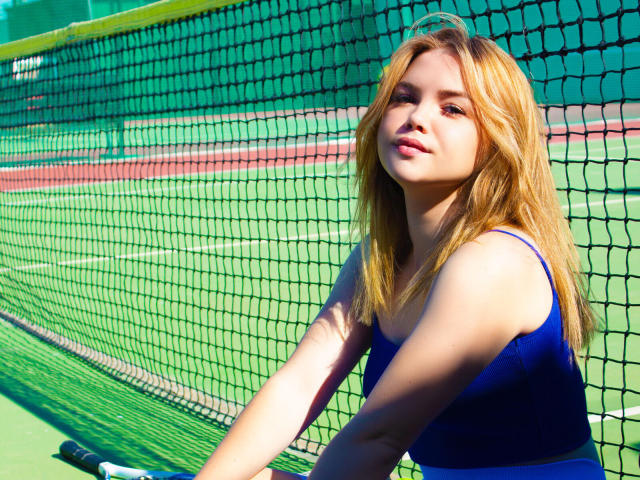 Kira Altman, com cabelo loiro, top esportivo azul, apoiada na rede de tênis, quadra verde, ensolarado, sorrindo, braços apoiados na rede, seios visíveis, magra, atraente, pele clara, atlética..