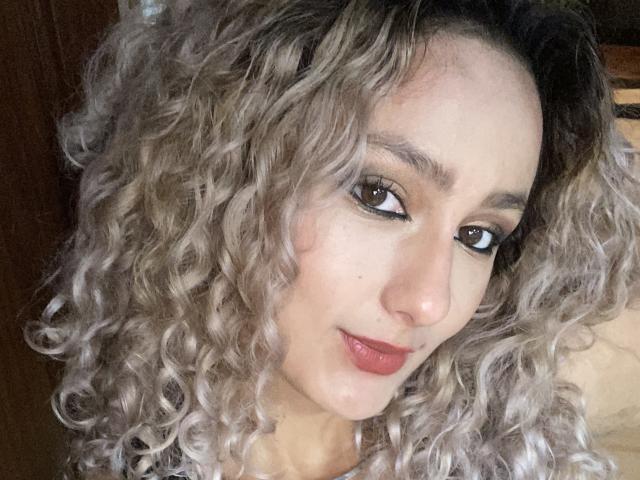 FloraSaenz has curly, grayish-blonde hair, brown eyes, and red lipstick. She's looking up with a slight smile. Her face is centered, and she's wearing a black top.