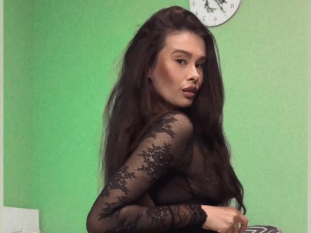 CameronMoss with long brown hair, wearing a black lace top, turning to look over her shoulder, showing her tits, in a green-walled room with a clock above.