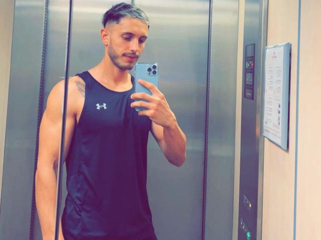 Muscular StevenCum with short, dyed hair, wearing a black Under Armour tank top, taking a mirror selfie in an elevator, showing a tattoo on his upper arm.