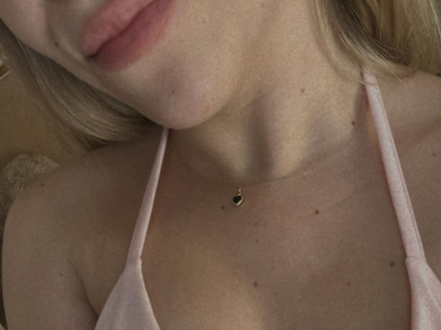 Le visage de SugarrX, montrant une partie de son haut rose à encolure américaine, sa peau claire, ses cheveux blonds et un petit collier en forme de cœur avec une pierre verte, près de ses seins..