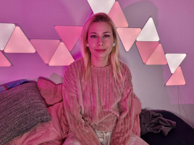 ViviVanilli-hot with blonde hair, pink ribbed sweater, and white pants, sitting on a bed, smiling, pink geometric wall lights, gray pillow, pink bed, and a black blanket.