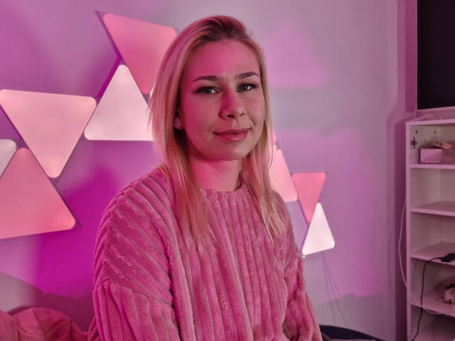 ViviVanilli – attraktiv mit blonden Haaren, einem pinken Pullover und einem leichten Lächeln, pinke Beleuchtung, weißer Hintergrund und dreieckigen Wanddekorationen, wirkt süß und entspannt..