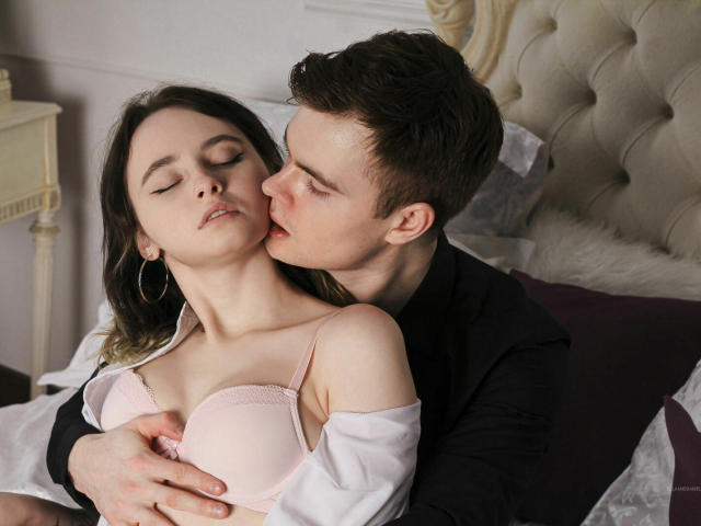 EllaAndDani, com os olhos fechados, vestindo um sutiã rosa, com os seios sendo delicadamente apertados por Dani, que tem cabelo castanho curto, uma camisa preta e está beijando o pescoço dela, na cama com uma cabeceira estofada..