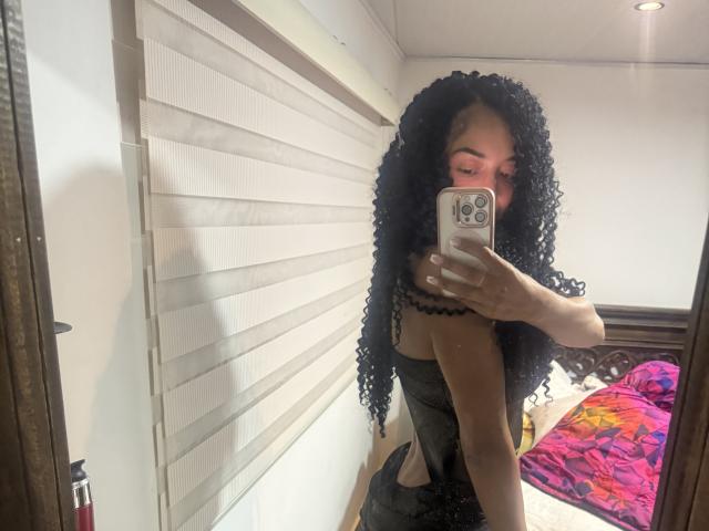 SofiColombiana, com cabelos pretos cacheados, vestindo uma blusa preta de renda, tirando uma selfie no espelho mostrando seu traseiro, segurando um iPhone rosa, em um quarto com uma cama colorida..