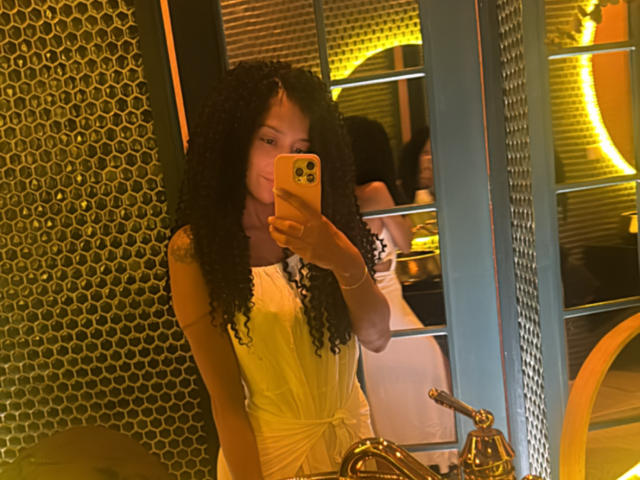 SofiColombiana met krullend zwart haar, gekleed in een gele jurk, maakt een spiegelzelfportret met een gele iPhone, waarbij haar kleine borsten en schattige gezicht te zien zijn..
