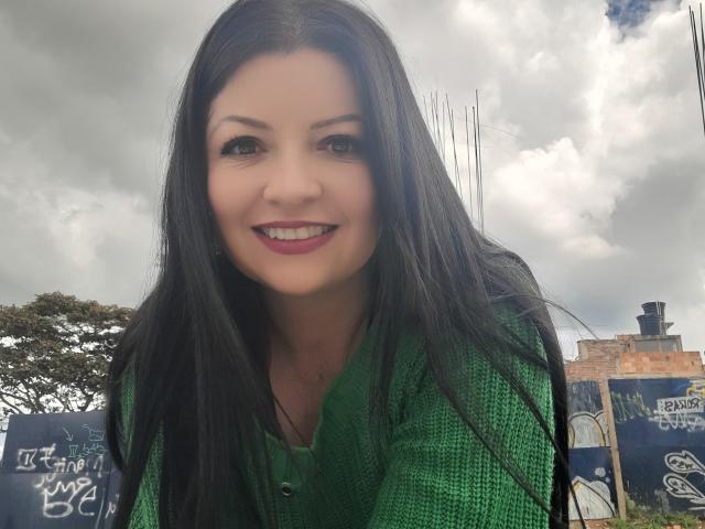 NatalyHami, com cabelos pretos longos e um suéter verde, sorrindo para a câmera, exibindo seus seios bonitos e batom rosa, com um céu nublado ao fundo..