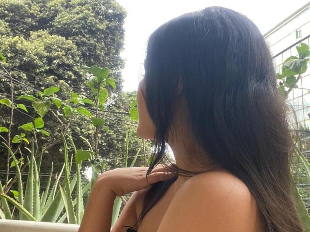 MiiaPalmer with long dark hair, facing left, topless, holding hair, visible ass, green leafy plants in background, sunny, outdoor.