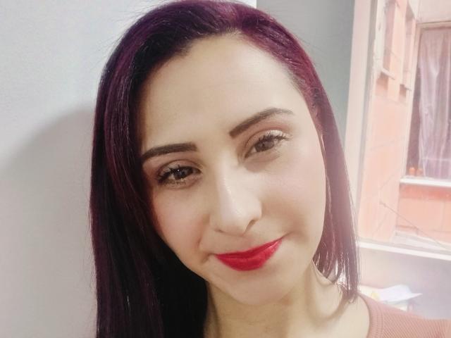 Karenma69 has dark purple hair, red lipstick, and brown eyes, smiling with a slight nose tilt, wearing a pinkish top, looking at the camera.