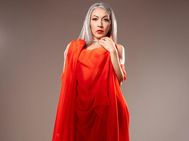 Nicolette Shea, con i suoi lunghi capelli argentati, il rossetto rosso e un abito rosso trasparente, lo tiene sopra il seno. Sta guardando la fotocamera con un'espressione seria..