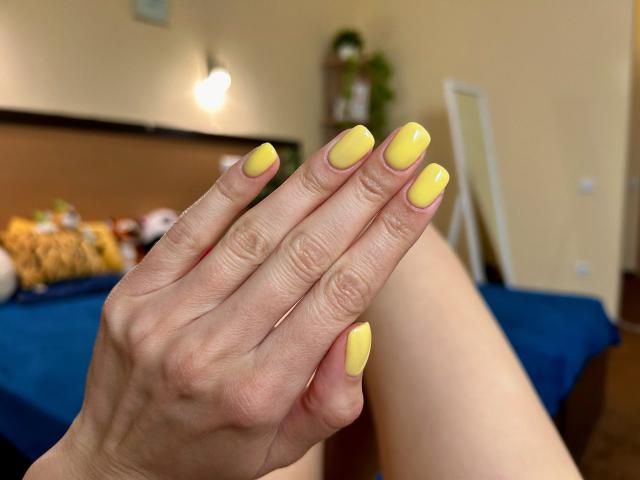 A mão de Karish, com unhas amarelas, brilhantes e quadradas, dedos abertos, exibindo unhas bem cuidadas e limpas. Sua pele é clara, e suas unhas são bonitas e bem mantidas..