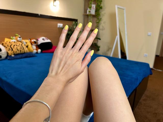 Karish's hand with yellow nails, fingers spread, rests on her bare thighs. She wears a silver bracelet, and her blue bed is visible with stuffed animals in the background.