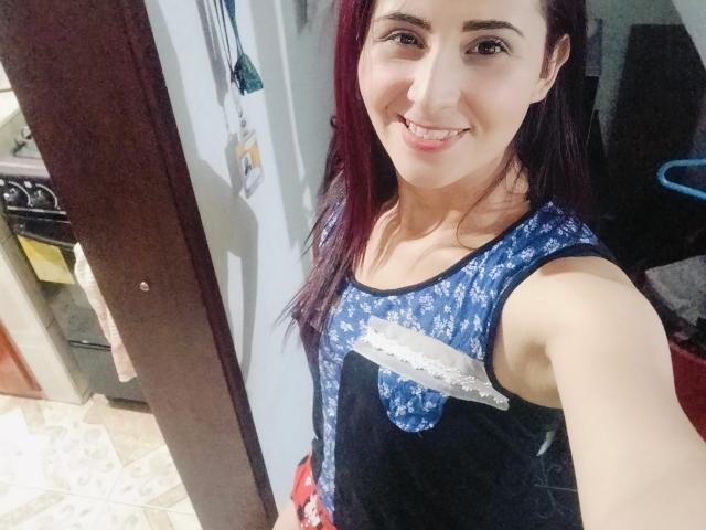 Karenma69 with dark red hair, smiling, wearing a blue and black floral tank top, showing her tits slightly, in a kitchen. She's holding a phone for a selfie.