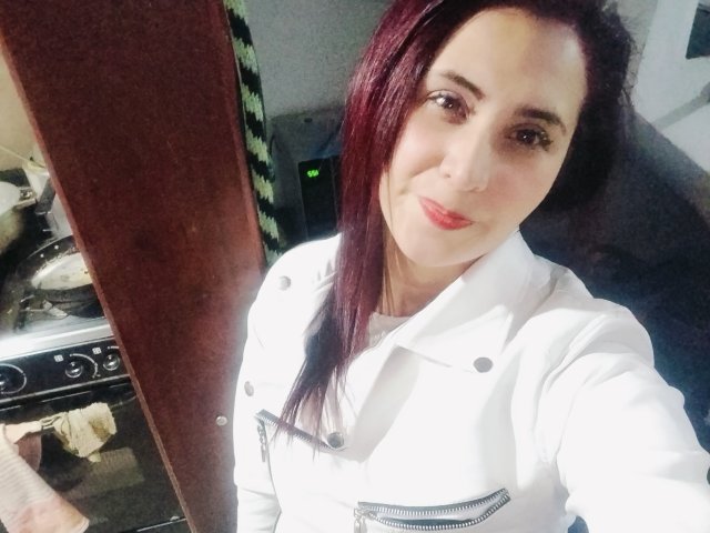 Karenma69 with red hair, wearing a white chef's coat, smiling at the camera, showing her tits slightly visible under the coat. She's holding a spoon in the left.