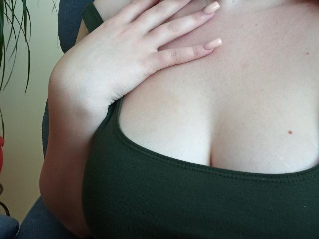 DiLaria's pale skin, showing her large tits in a green tank top, with her hand gently touching her chest. Small mole on her right breast.