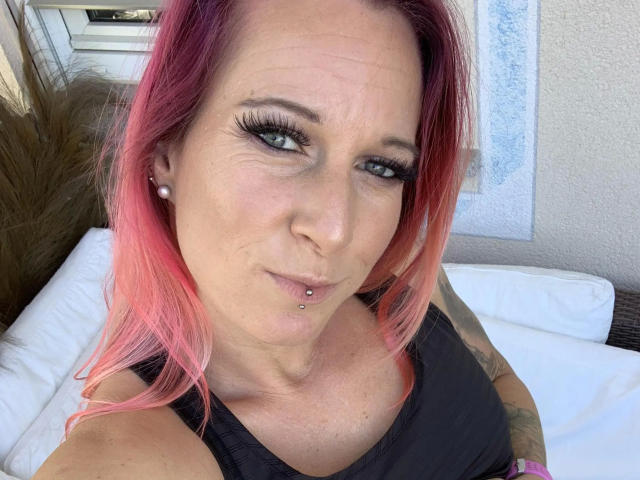 Candy-bitch-hot with pink-dyed hair, black tank top, and tattooed arm, sporting a lip piercing and long eyelashes, looks at the camera with a slight smirk.