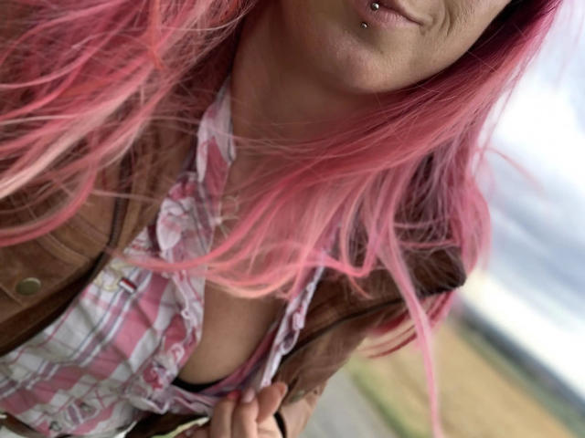 Candy-bitch-hot with pink hair, pierced lips, wearing a brown vest and pink checkered shirt, exposing a small bit of her tits, holding the shirt.