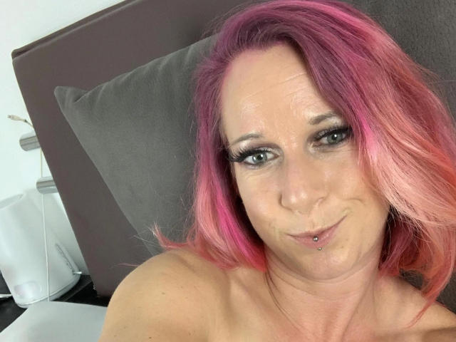 Candy-bitch-hot with pink hair, green eyes, and a pierced lower lip, smiling at the camera. She's topless, showing her bare shoulders. Her tits are slightly visible.