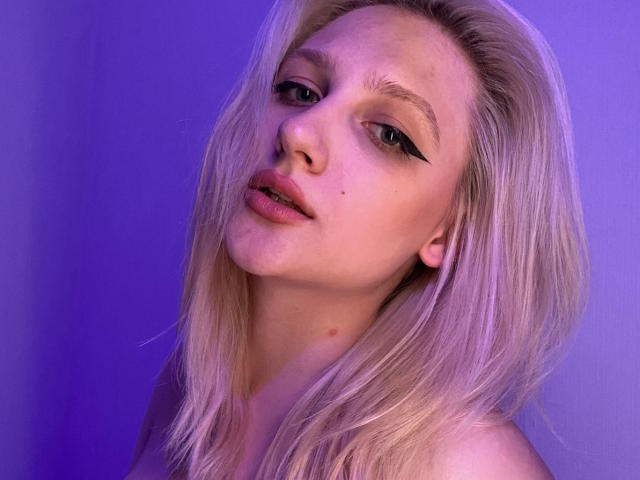 Blond Xloxy con maquillaje ligero, delineador de ojos negro grueso, labios rosados ligeramente separados y una pequeña peca en su mejilla izquierda, mirando a la cámara con una expresión suave..