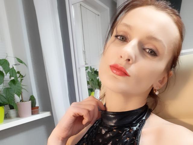 Sympathy heeft glanzende rode lippen, een lichte huid, bruine ogen en draagt een zwart leren topje met halter. Ze raakt haar nek aan met één hand en kijkt naar de camera..