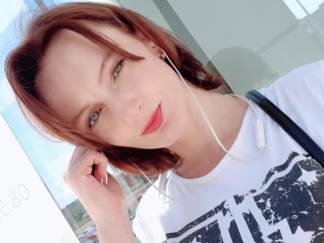 Medeleven met rood haar, witte oordopjes en een lichtjes grijnzend gezicht, gekleed in een wit shirt met zwarte tekst. Hand in de buurt van het oor, lichtroze lippenstift..