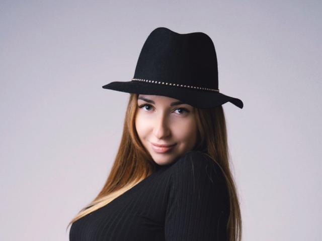 AriaGorgeous with long brown hair, black hat, and black ribbed top, showing a slight smile and looking over her shoulder, has small tits and a cute face.