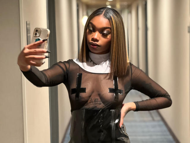 ItzAphroditee tomando un selfie en un pasillo, vistiendo una blusa negra transparente con cubrebustos negros con forma de cruz, sosteniendo un teléfono, y con una falda de cuero negro..