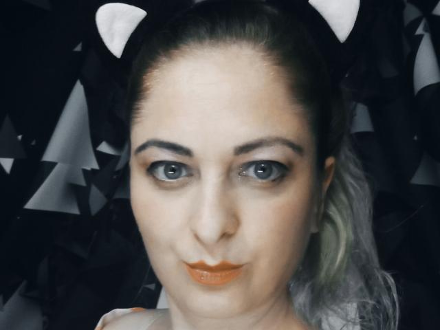 MonicaMiss has orange lipstick, grey eyes, and white cat ears on a black headband. Her eyebrows are thick and she's looking directly at the camera with a slight smile.