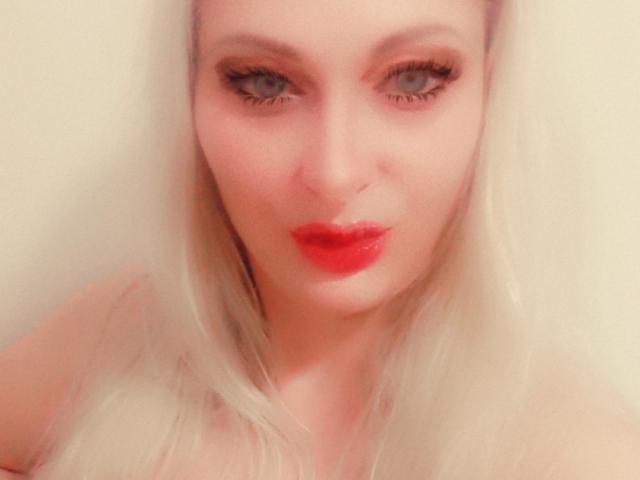 MonicaMiss has pale skin, long white hair, red lipstick, and dark eyeliner. Her blue eyes are slightly squinted, and she's giving a pouty, almost kissy, facial expression.