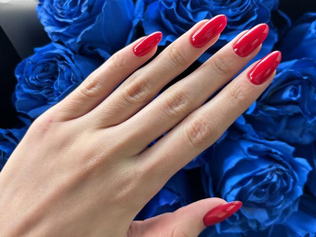 Pingli's hand with shiny red nails, showcasing long, pointed tips, rests against a cluster of blue roses. Her skin is light and smooth.