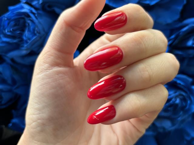 Pingli's shiny red nail polish covers her long, glossy nails on a pale hand. She's holding it up against a blue rose background.