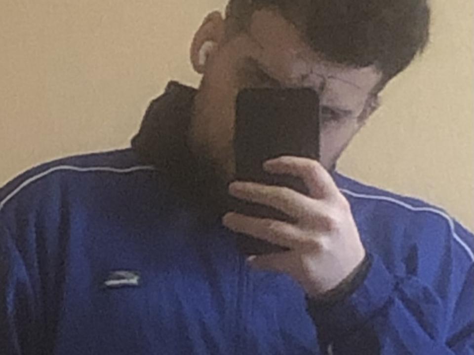 Yepzor taking a mirror selfie, wearing a blue Adidas track jacket with white stripes, holding a black phone, short dark hair, serious expression, tan skin, slightly blurry.